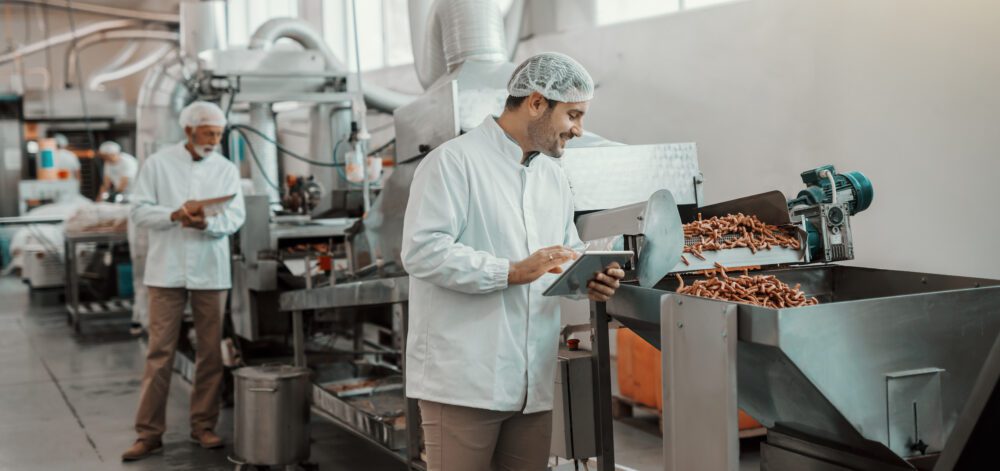 Young Caucasian supervisor evaluating quality of food in food plant while holding tablet. Man is dressed in white uniform and having hair net.